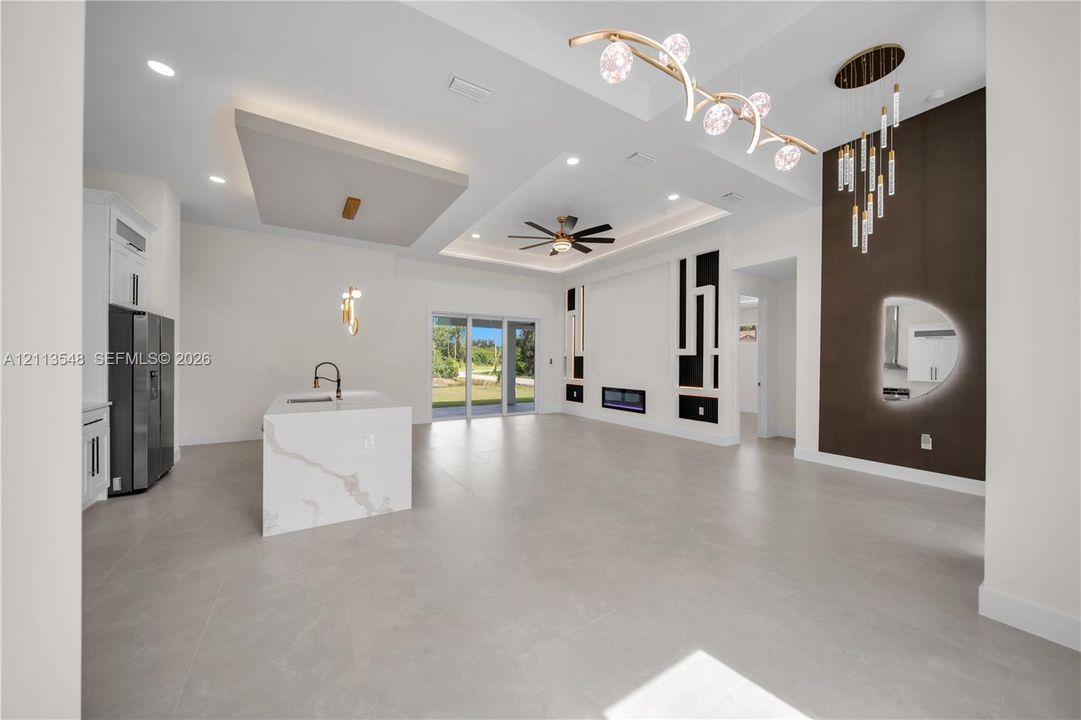 Open-concept interior featuring large-format tile flooring, a kitchen island with a stone-surfaced countertop, a tray ceiling with recessed lighting, an integrated fireplace, and decorative wall paneling