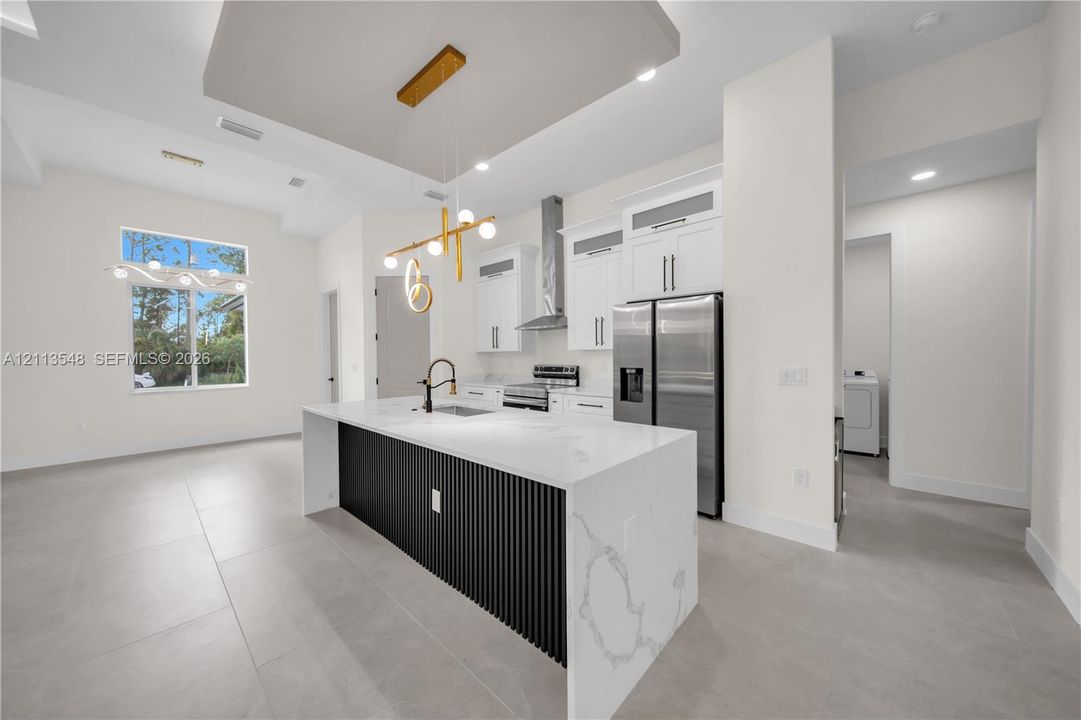 Kitchen featuring a central island with a stone-finish countertop and vertical slat detailing, white shaker-style cabinetry, stainless steel appliances, a modern multi-light pendant fixture, and large-format tile flooring