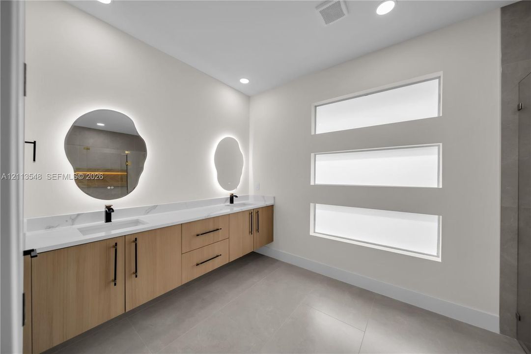 Floating vanity featuring wood-finish cabinetry, solid surface countertop with integrated dual sinks, matte black hardware, backlit organic-shaped mirrors, and horizontal transom windows