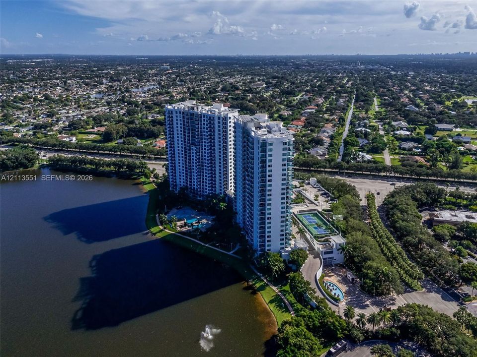 High-rise residential development featuring a multi-story glass-facade tower, ground-level swimming pool, tennis courts, and waterfront positioning on a large lake
