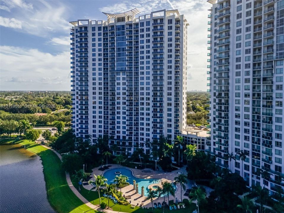 High-rise residential exterior featuring expansive glass balconies, a resort-style swimming pool, integrated spa, and manicured waterfront grounds
