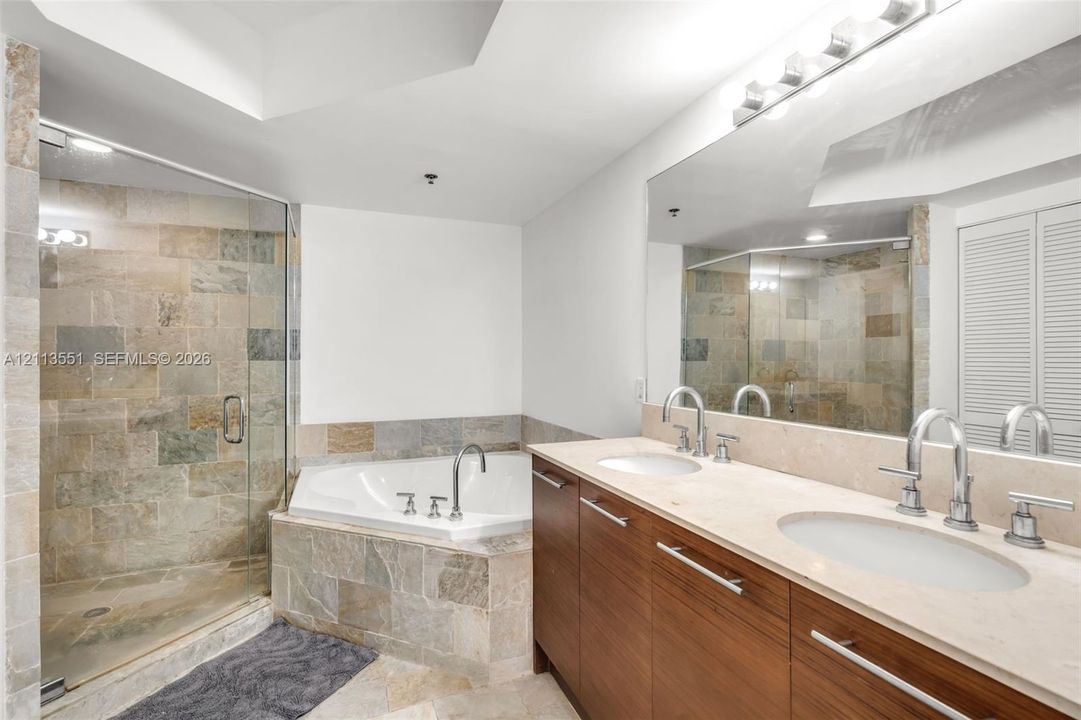 Bathroom featuring a dual-sink vanity with wood-finish cabinetry and light-toned stone countertop, a glass-enclosed shower with textured stone tile, and a corner soaking tub with stone tile surround