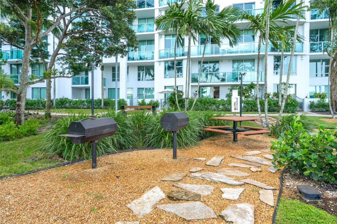 Outdoor common area featuring built-in charcoal grills, stone paver pathways, integrated picnic table, and mature tropical landscaping