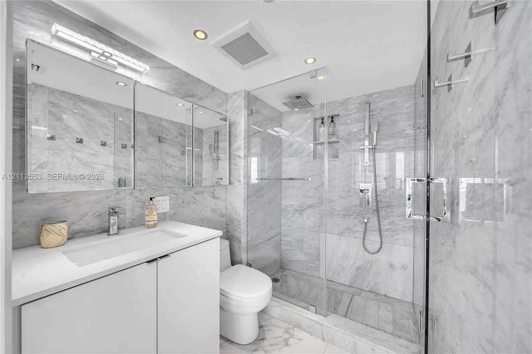 Bathroom featuring wall-to-wall natural stone tiling, a glass-enclosed walk-in shower with integrated shelving, a modern vanity with a white countertop, and a recessed medicine cabinet with integrated lighting