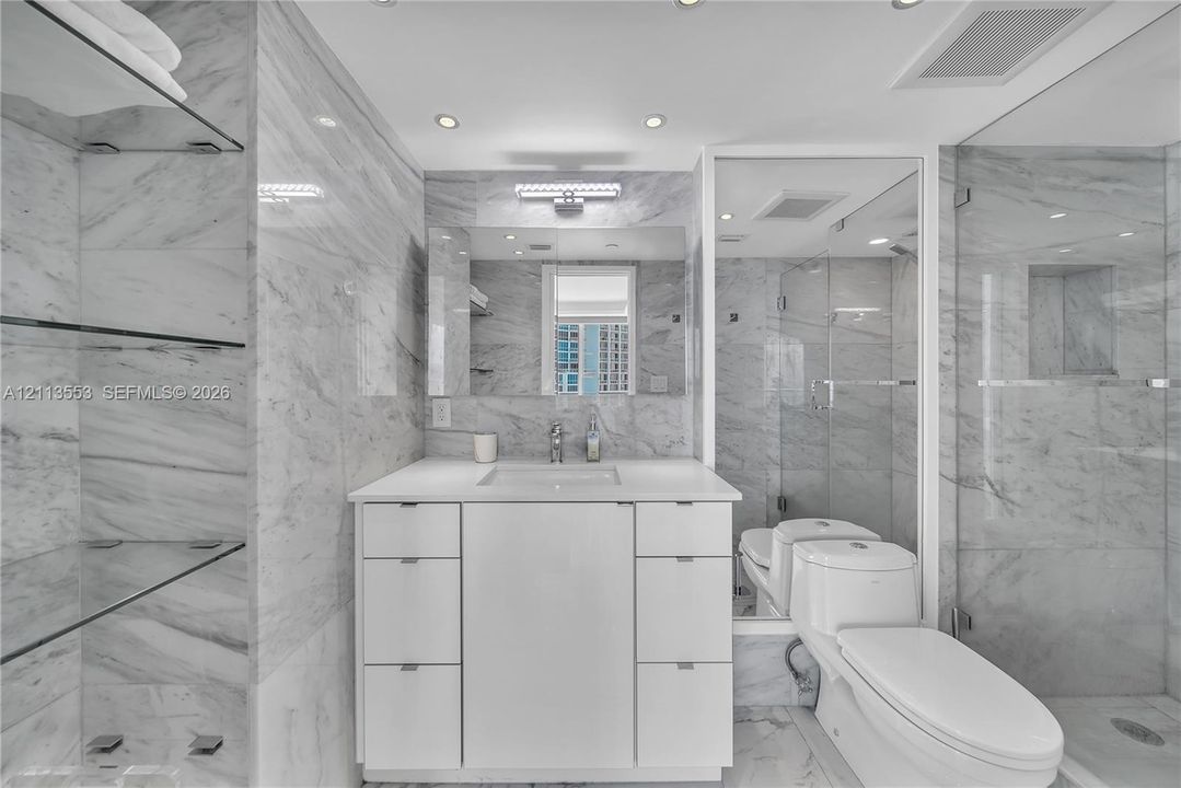 Bathroom featuring floor-to-ceiling stone-tiled walls, a white vanity with integrated basin, recessed lighting, glass shelving, and a frameless glass-enclosed shower