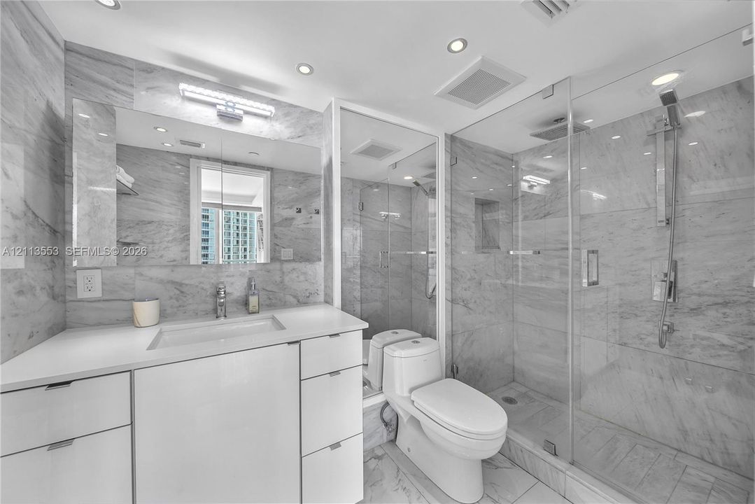 Bathroom featuring large-format stone wall tiling, white cabinetry with a solid-surface countertop, glass-enclosed walk-in shower with rainfall showerhead, and recessed ceiling lighting