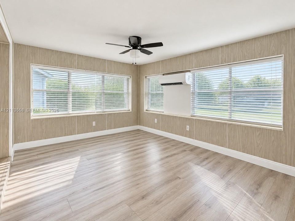 Interior space featuring wood-finish flooring, wood-panel walls, multiple windows with integrated blinds, a ceiling fan, and a wall-mounted climate control unit