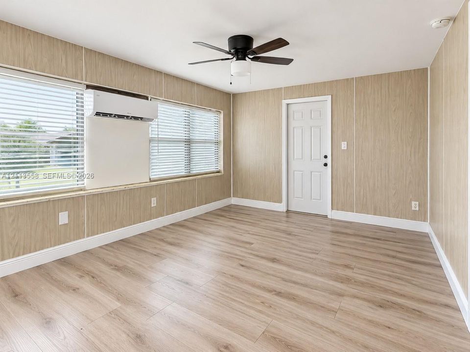 Room featuring wood-finish flooring, paneled walls, a ceiling fan with integrated lighting, a wall-mounted ductless air conditioning unit, and multiple windows with horizontal blinds