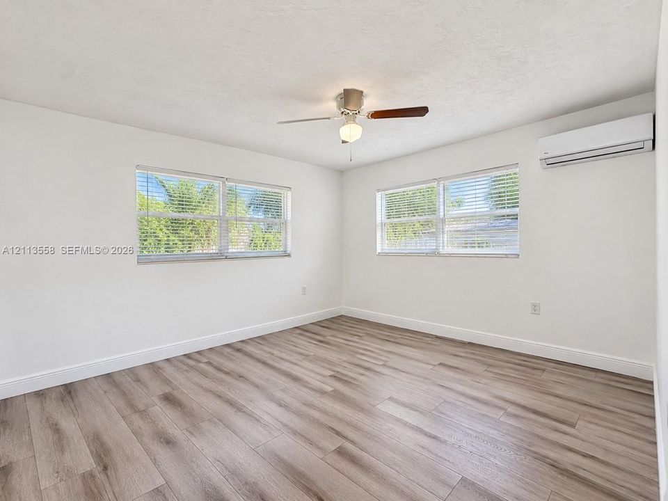 Wood-finish flooring, dual windows with horizontal blinds, ceiling fan with integrated lighting, and wall-mounted ductless climate control unit