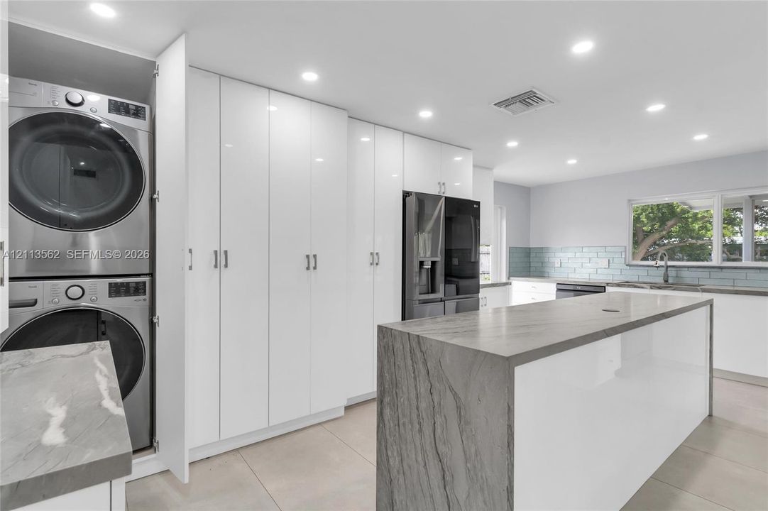 Kitchen featuring a waterfall-edge island with stone-finish countertop, high-gloss white cabinetry, integrated laundry station, and recessed lighting