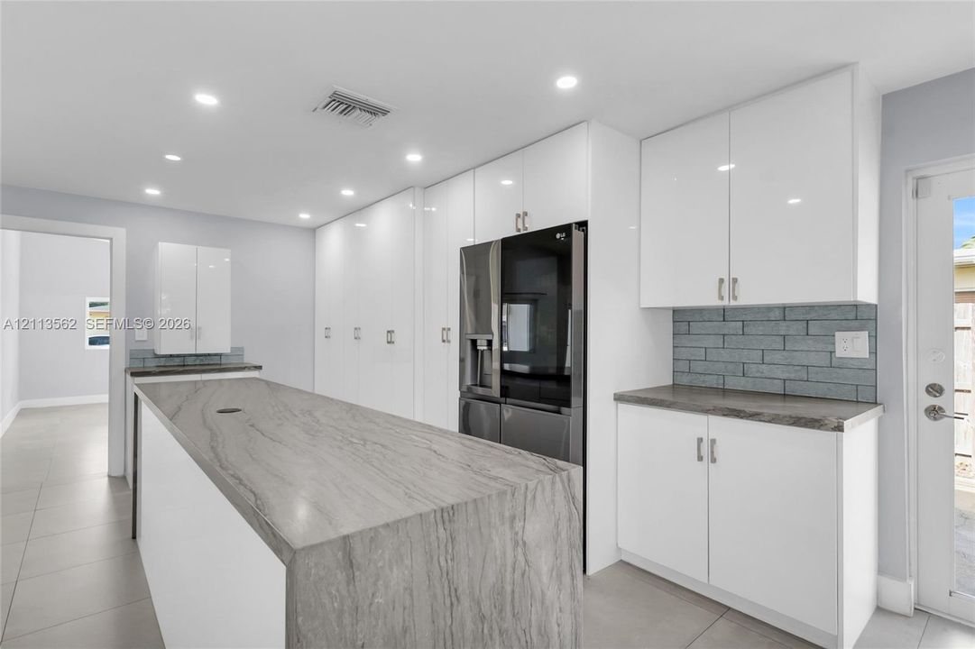 Kitchen featuring white flat-panel cabinetry, stone-surface waterfall island, grey brick-pattern backsplash, and tile flooring