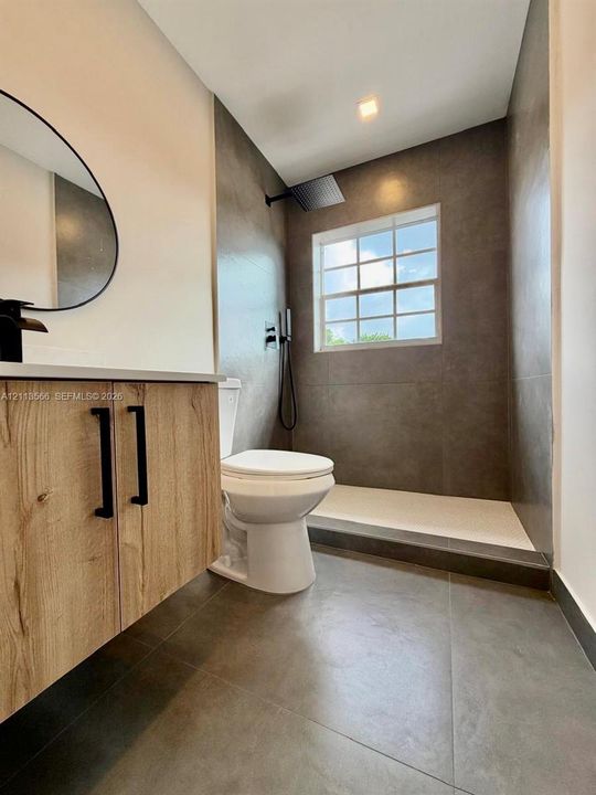 Bathroom featuring a wood-finish vanity, large-format grey tile flooring, floor-to-ceiling tiled shower surround, matte black hardware, and an oval wall-mounted mirror