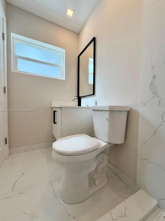 Bathroom featuring white stone-finish flooring and wall tile with grey veining, a white vanity with a stone countertop, a rectangular mirror with a black frame, and a matte black faucet