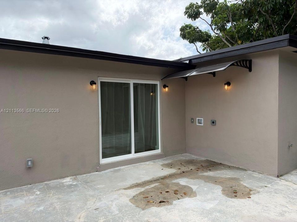 Concrete patio featuring sliding glass doors, wall-mounted exterior lighting, and a metal door awning