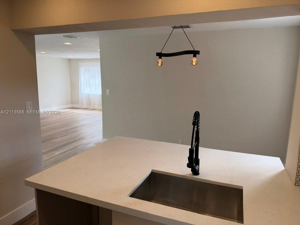 Kitchen island featuring a solid-surface countertop, an undermount rectangular stainless steel sink, and a matte black gooseneck faucet with a pull-down sprayer, complemented by a suspended modern geometric lighting fixture and wood-finish flooring