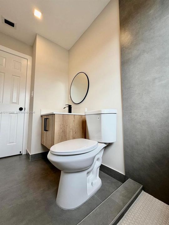 Floating wood-finish vanity with a stone-top counter, matte black faucet, oval wall-mounted mirror, and large-format gray tile flooring