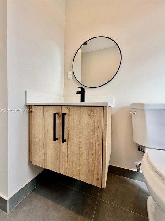 Wall-mounted vanity featuring wood-finish cabinetry, a white countertop, matte black hardware, and a circular mirror with a black frame