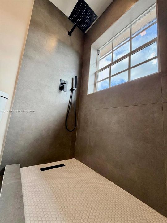 Walk-in shower featuring large-format wall tile, a matte black rainfall showerhead, a handheld sprayer, a multi-pane window, and a penny-round tile floor with a linear drain