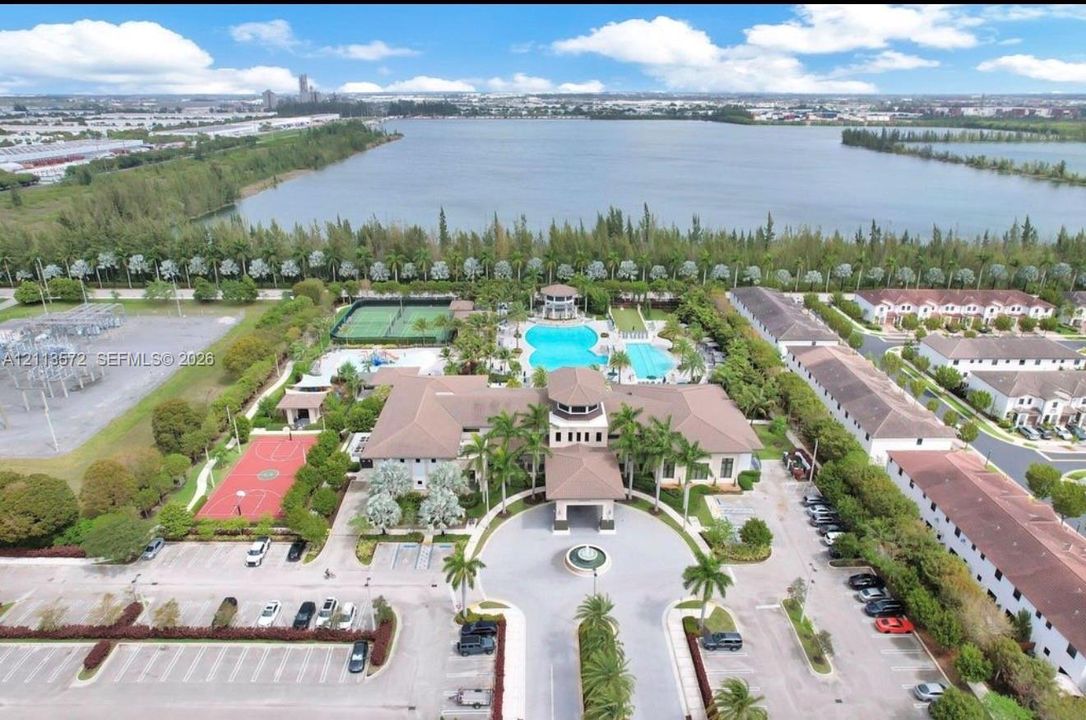 Aerial perspective of the community center featuring a swimming pool, tennis court, basketball court, and central fountain, situated alongside a lake
