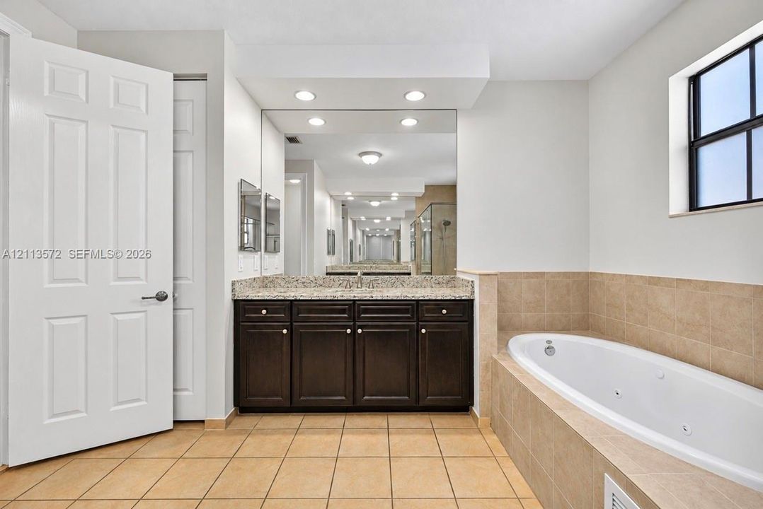 Bathroom featuring a dark-finish vanity, stone-pattern countertop, recessed lighting, wall-to-wall mirror, and a jetted soaking tub with tile surround