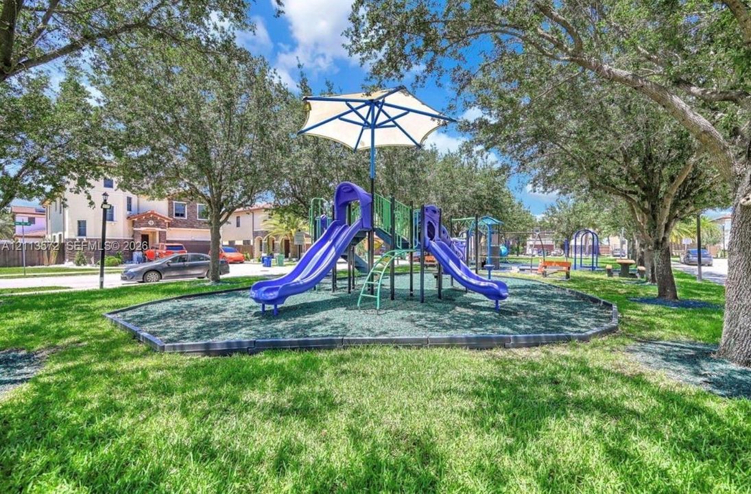 Community park featuring a central playground structure with dual slides, a canopy cover, manicured lawn, and mature canopy trees