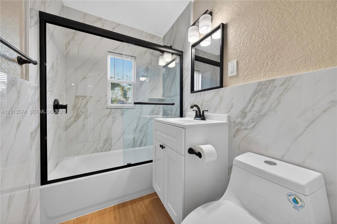 Bathroom featuring a tub-shower combination with floor-to-ceiling stone-style tile surround and a black-framed sliding glass door, white vanity with a rectangular sink, matte black hardware, and wood-finish flooring