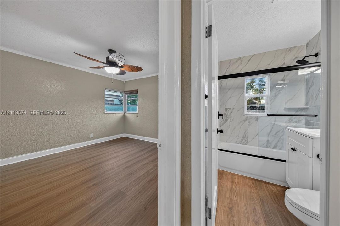 Room featuring wood-finish flooring and a ceiling fan, paired with a bathroom featuring stone-pattern wall tile, a glass-enclosed shower, and a white vanity