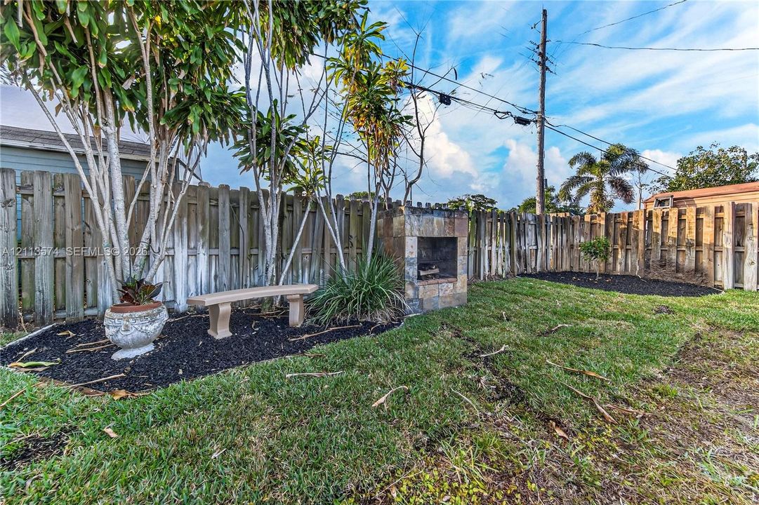 Rear yard featuring a stone-clad outdoor fireplace, perimeter wood fencing, dark mulch garden beds, and mature landscaping