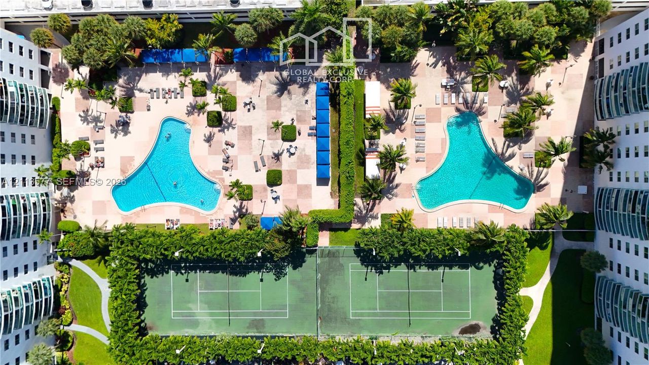 Recreational area featuring two outdoor swimming pools, two hard-court tennis courts, paved decking, and mature landscaping