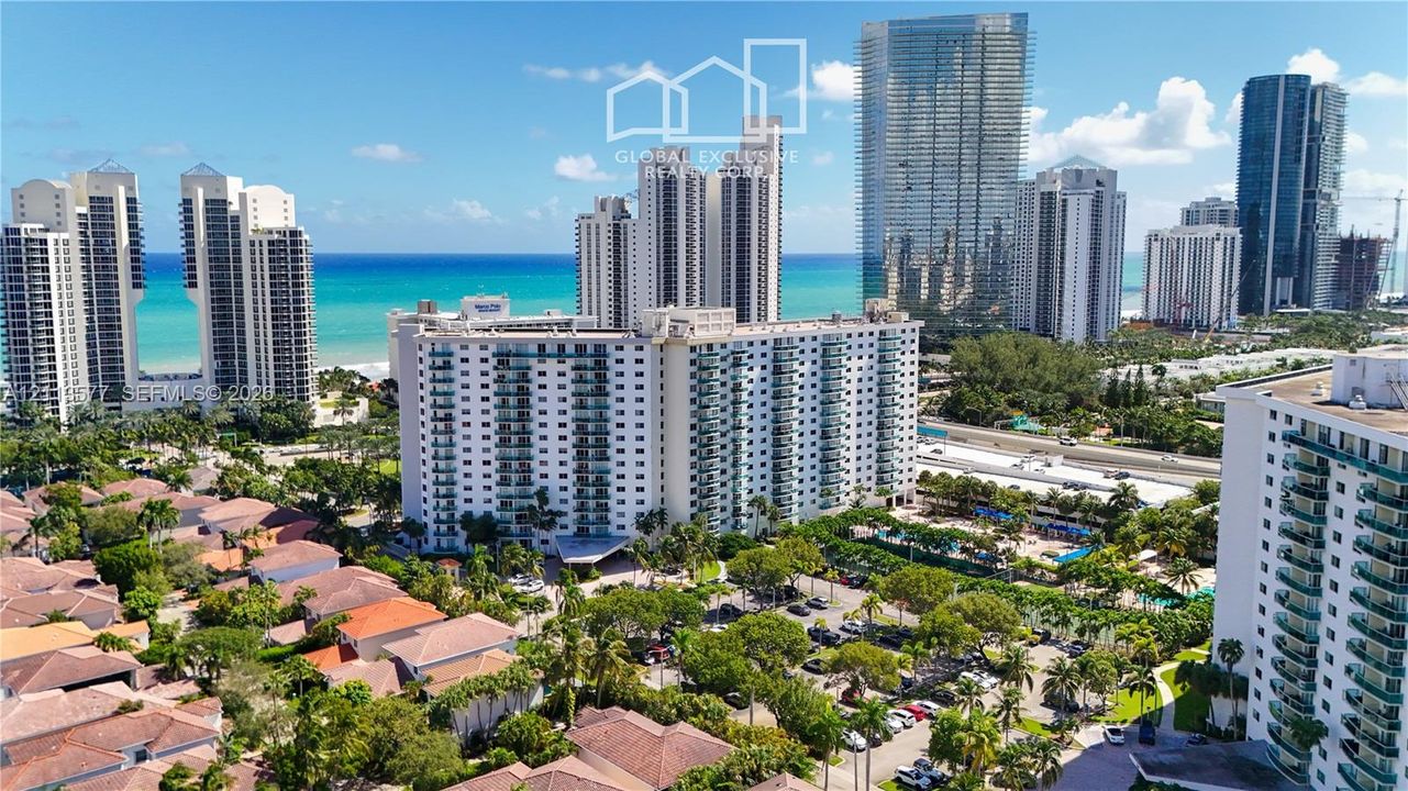 Aerial perspective featuring a multi-story residential building with private balconies, surrounded by mature palm trees, paved parking areas, and proximity to the coastline