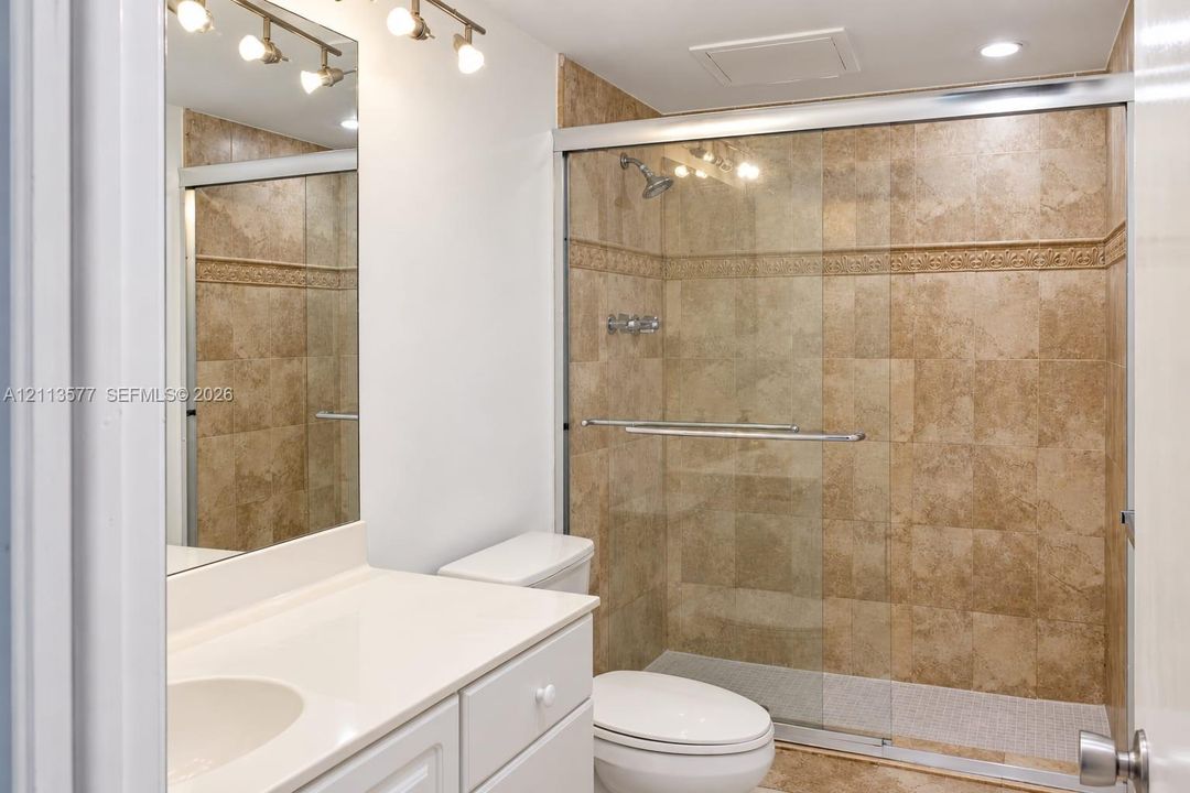 Bathroom featuring a vanity with a white countertop, an integrated sink, a full-length wall mirror, multi-light fixture, and a shower enclosure with floor-to-ceiling stone-finish tile surround and sliding glass doors
