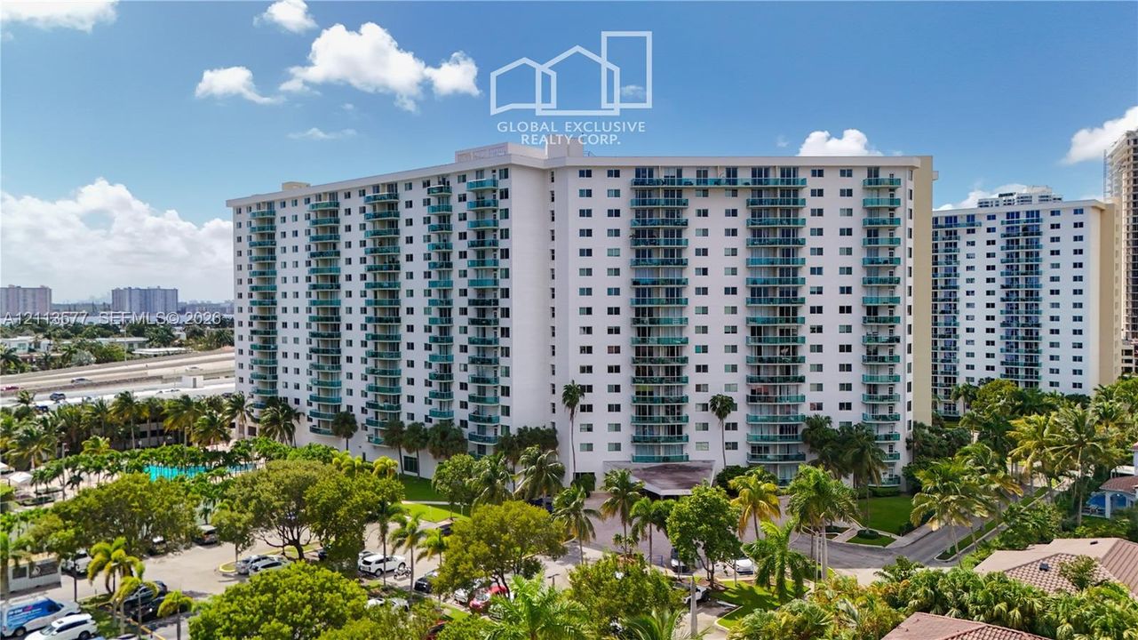 High-rise residential building featuring stacked private balconies with glass railings, an exterior swimming pool, mature palm tree landscaping, and paved parking areas