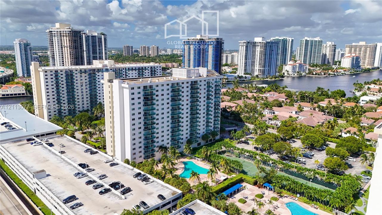 Aerial perspective of a multi-story residential complex featuring an outdoor swimming pool, tennis courts, paved parking structures, and mature landscaping