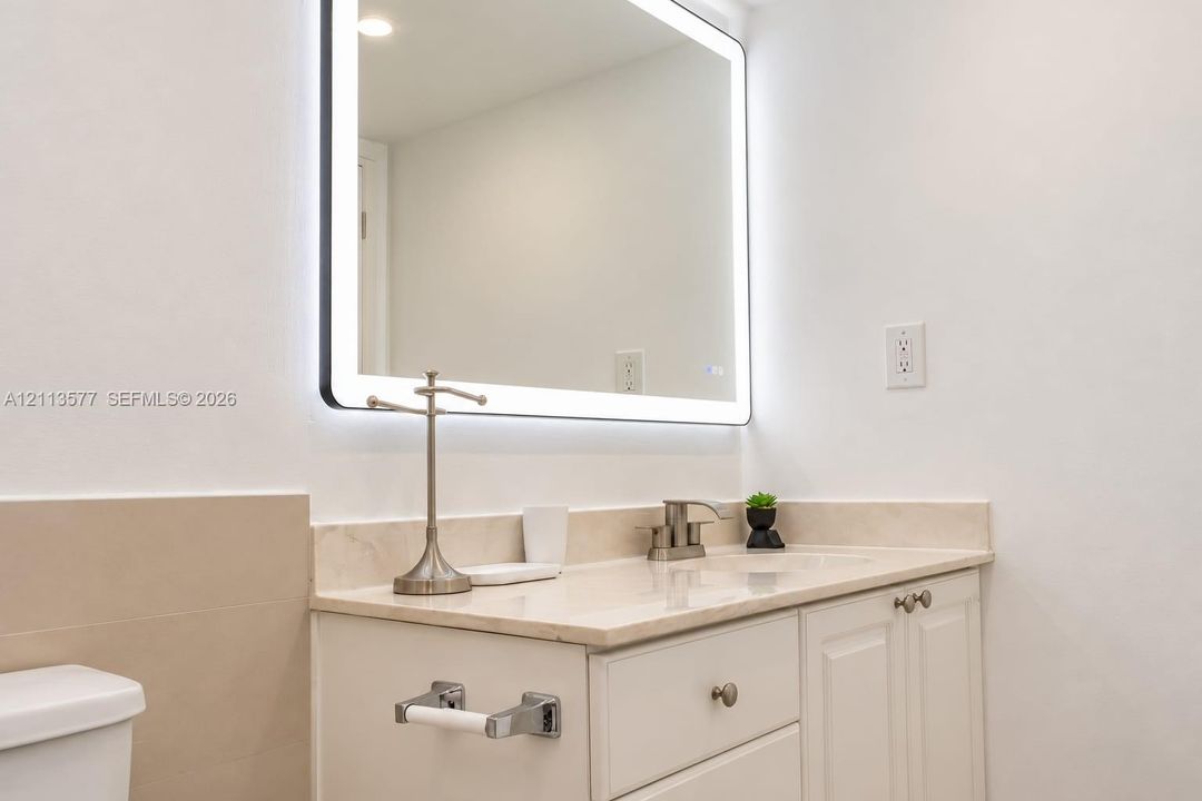 Rectangular LED-backlit mirror featuring rounded corners, paired with a white vanity cabinet, solid-surface countertop with integrated basin, and brushed-finish faucet