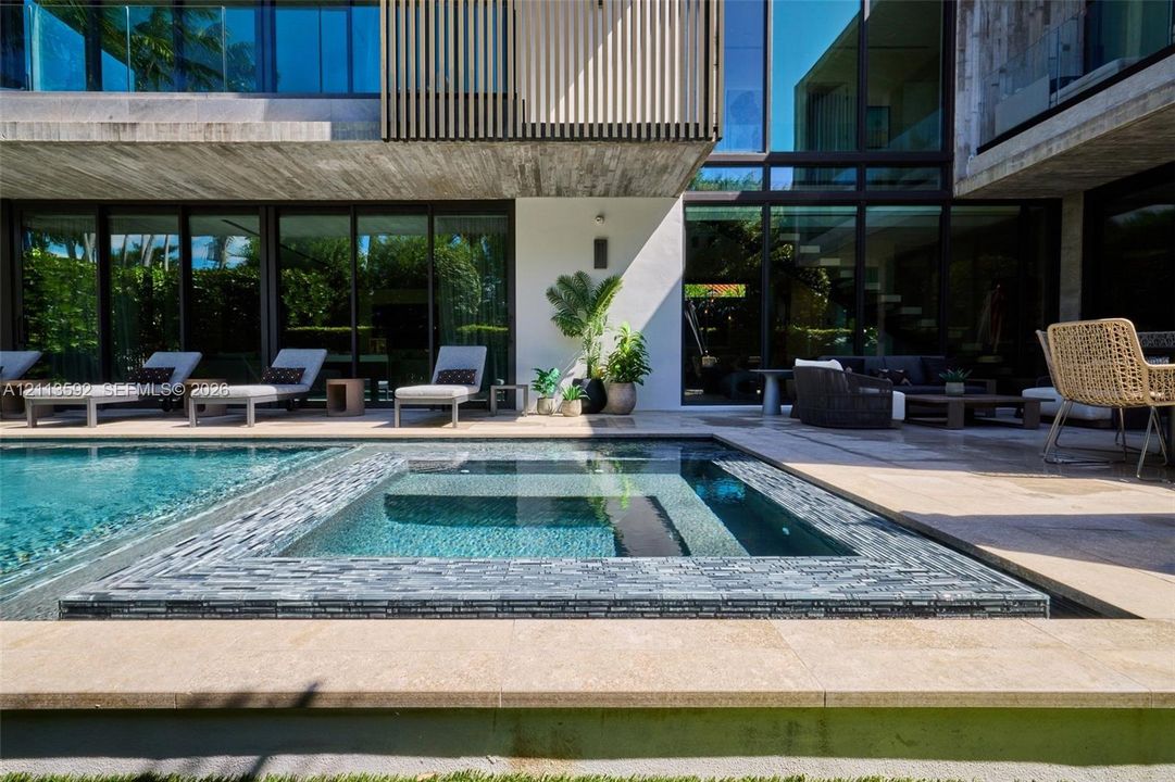 Modern exterior architecture featuring board-formed concrete walls, floor-to-ceiling glass sliding doors, custom wood-slat screening, and a sunken pool and spa with decorative stone tiling, set within a paved stone patio area