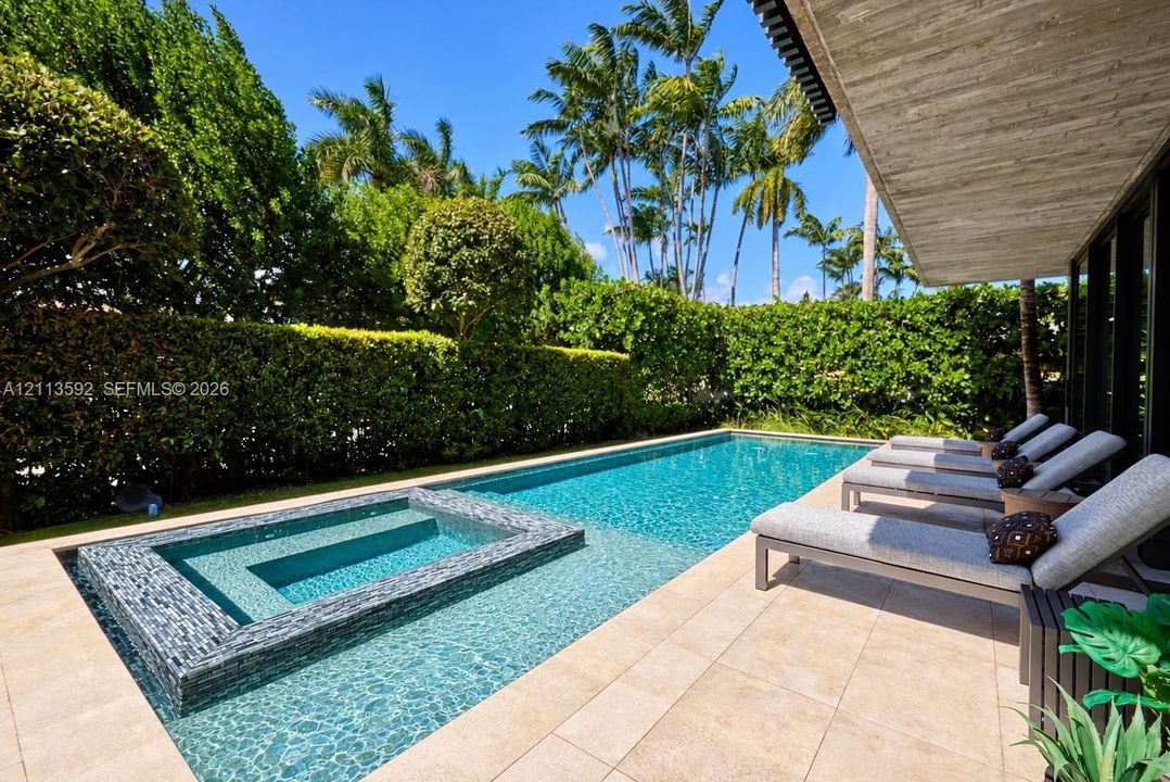 Outdoor living area featuring a custom swimming pool, integrated spa with mosaic tile accents, light-toned stone decking, and mature perimeter landscaping
