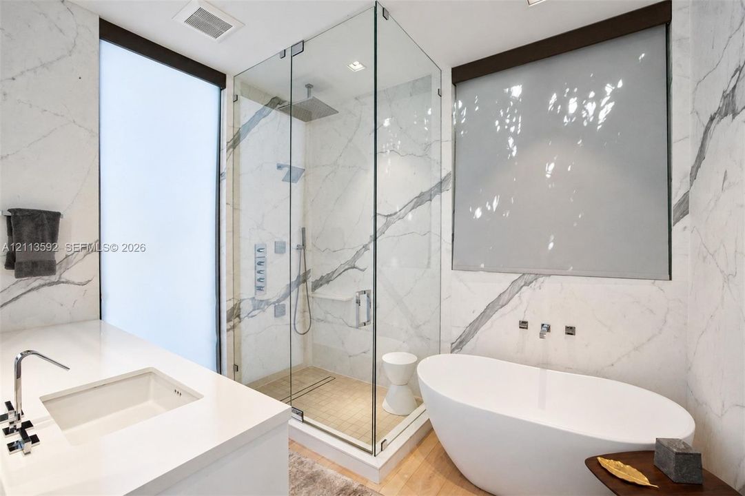 Bathroom featuring white veined wall paneling, a glass-enclosed shower with integrated body sprays and a ceiling-mounted rainfall showerhead, a freestanding soaking tub with wall-mounted faucet, and a vanity with a rectangular undermount sink