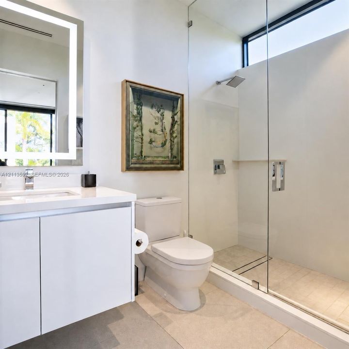 Modern white cabinetry with integrated countertop, backlit rectangular vanity mirror, and glass-enclosed shower with wall-mounted rainfall fixture and horizontal clerestory window