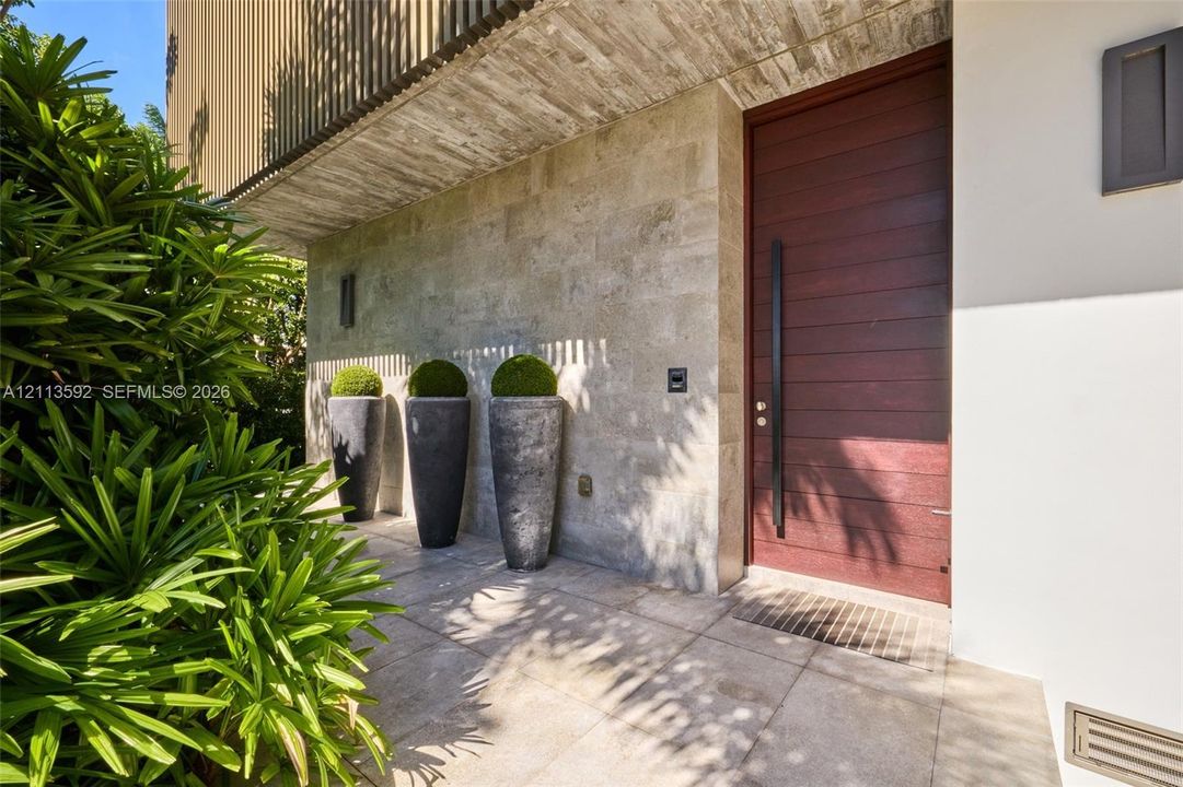 Entryway featuring a solid wood door with a vertical metal handle, natural stone wall cladding, a concrete ceiling overhang, large format floor tiling, and integrated wall sconce lighting
