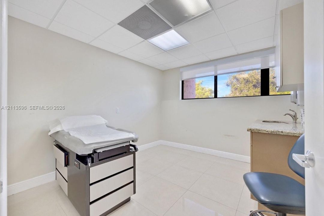 Drop-ceiling with integrated lighting, large window with roller shade, neutral tile flooring, stone-surfaced vanity with sink, and upper cabinetry