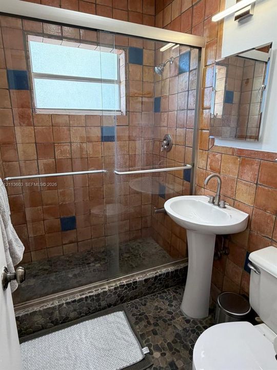 Bathroom featuring a walk-in shower with a sliding glass door, terracotta-style wall tile with navy blue accents, a pedestal sink, and pebble-stone flooring