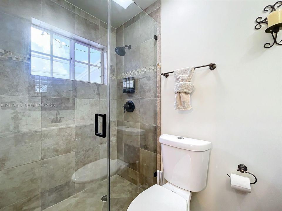 Walk-in shower with stone-tile surround, glass enclosure, and matte black hardware, accompanied by a pedestal-style toilet and wall-mounted light fixture