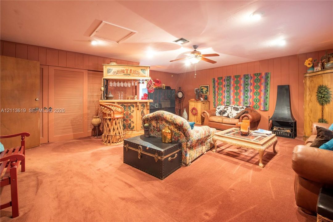 Wall-mounted metal fireplace, built-in wooden bar, wood-paneled walls, and ceiling fan with integrated lighting