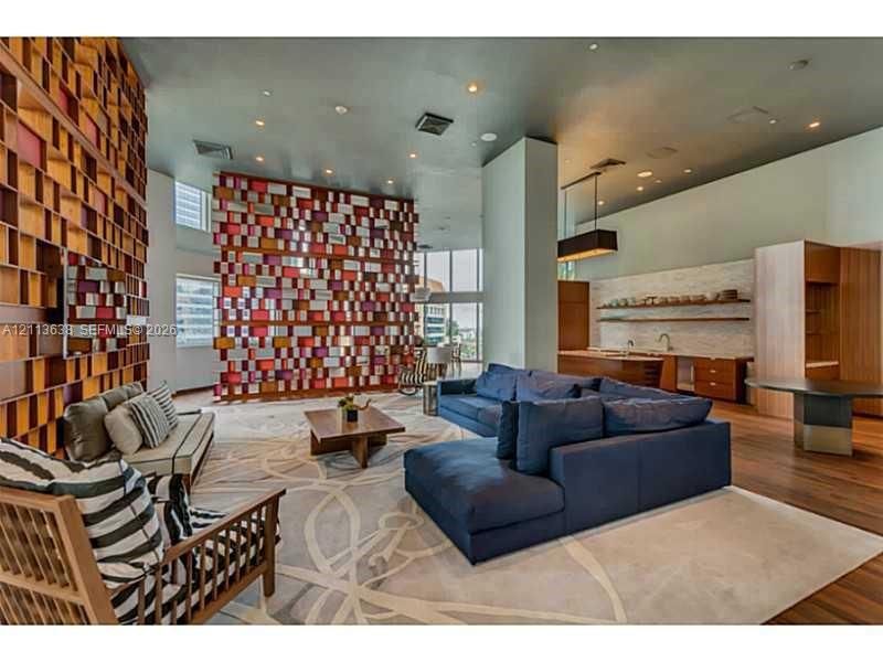 Open-concept living space featuring high ceilings, a prominent floor-to-ceiling geometric wood-toned feature wall, wood-finish flooring, and a built-in wet bar with wood cabinetry and open shelving