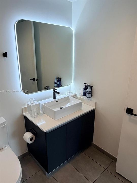 Modern vanity featuring a rectangular vessel sink, white stone-finish countertop, dark cabinetry, and a wall-mounted matte black faucet, accented by a backlit rounded mirror