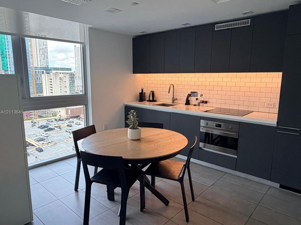 Modern kitchen featuring matte charcoal cabinetry, light-toned stone-finish countertops, a white subway tile backsplash with under-cabinet lighting, a built-in stainless steel oven, and floor-to-ceiling windows