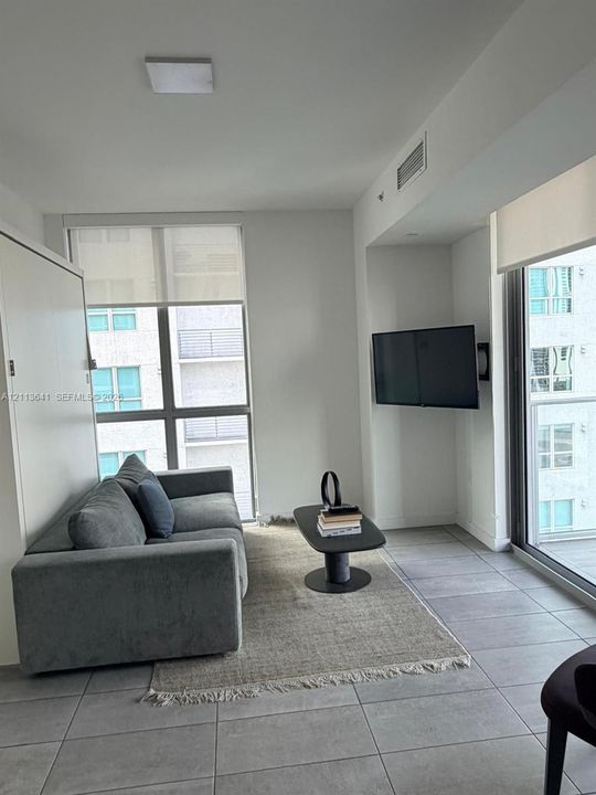 Floor-to-ceiling windows with roller shades, neutral tile flooring, built-in Murphy bed cabinetry, and wall-mounted television bracket