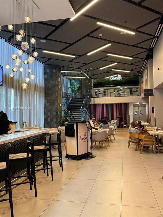 Interior space featuring floor-to-ceiling windows, tiered globe pendant lighting, stone-tiled support columns, grid-patterned dropped ceiling with integrated linear lighting, and a staircase with metal railing