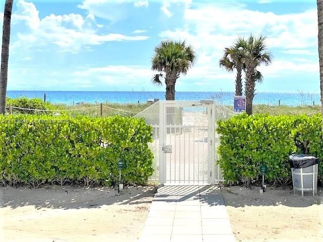 Sand-colored paver walkway leading to a white metal security gate flanked by manicured privacy hedges and palm trees