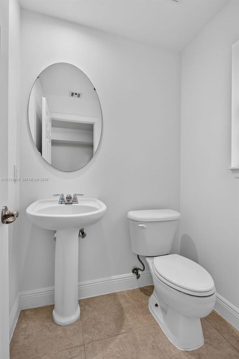 Ceramic tile flooring, white pedestal sink with chrome faucet, oval wall-mounted mirror, and standard porcelain toilet